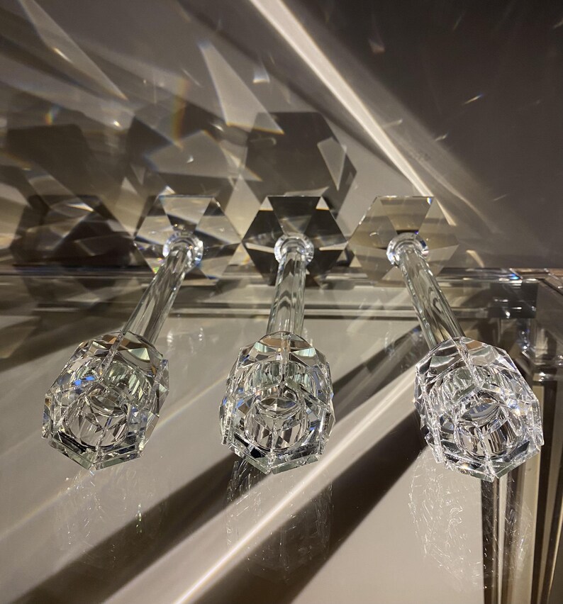 K&ouml;nnte beinhalten: Drei Kerzenhalter aus klarem Glas mit facettiertem, sechseckigem Design. Jeder Halter verf&uuml;gt &uuml;ber einen hohen, schlanken Stiel und einen breiten, geometrischen Fu&szlig;, der das Licht reflektiert und scharfe Schatten sowie helle Lichtpunkte erzeugt.