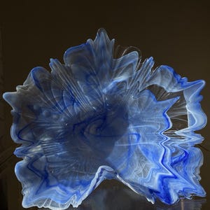 May include: A decorative glass bowl featuring a swirling pattern of deep blue and translucent white colours. The bowl has an organic, irregular shape with ruffled, wavy edges that resemble the form of a large, open leaf or flower petal.