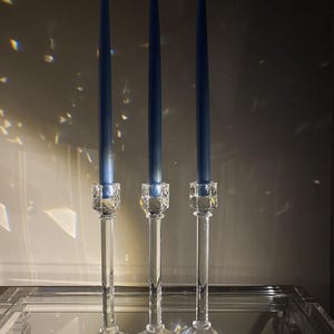 K&ouml;nnte beinhalten: Drei hohe, schlanke, dunkelblaue Stabkerzen stehen aufrecht in klaren, facettierten Kristallkerzenhaltern. Jeder Halter verf&uuml;gt &uuml;ber einen sechseckigen Fu&szlig;, einen glatten zylindrischen Schaft und ein strukturiertes, geometrisches Oberteil.