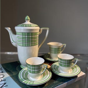 May include: A vintage tea set featuring a white teapot, creamer, and two teacups with saucers. The set has a green plaid pattern and green accents. The teacups and saucers are on a book.