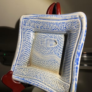 May include: A square ceramic dish featuring a white base with blue textured edges and a central design of embossed wavy lines and a small comet-like shape. The surface has a speckled finish with a distinct blue border and a smooth, slightly concave center.