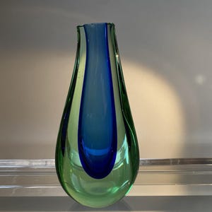 May include: A teardrop-shaped glass vase with a vibrant colour scheme. The vase features a central column of deep blue, framed by a lighter blue, and encased in a translucent green exterior. The vase is displayed on a clear glass shelf.