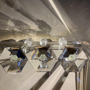 K&ouml;nnte beinhalten: Drei klare Glaskerzenhalter in einer Reihe, jeder mit einem sechseckigen Fu&szlig; mit abgeschr&auml;gten Kanten und einem facettierten Schaft. Das transparente Material reflektiert das Licht und erzeugt scharfe geometrische Schatten und helle Lichtpunkte.