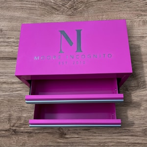 May include: A magenta-colored box with two pull-out drawers. The top of the box features the letter "N" and the words "MOORE INCOGNITO EST. 2010" in a lighter color. The drawers have light blue accents.