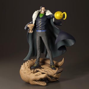 Op de afbeelding: Gedetailleerd beeldje van een personage uit de anime-serie One Piece. Het beeldje staat op een getextureerde basis met een krokodillenontwerp. Het personage draagt een donkere jas, een groen overhemd en houdt een gouden haak en een bruin voorwerp vast.