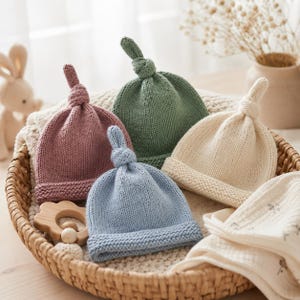 Könnte beinhalten: Vier gestrickte Babymützen mit Knoten an der Spitze in Altrosa, Salbeigrün, Creme und Hellblau. Jede Mütze hat einen umgeschlagenen Rand im Kraus-Rechts-Muster und eine glatt rechts gestrickte Krone. Sie liegen zusammen in einem Weidenkorb.