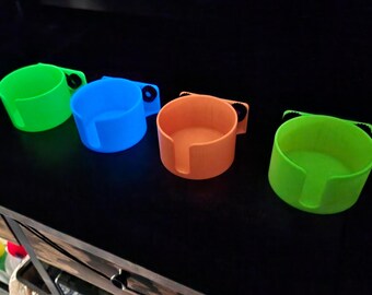 Könnte beinhalten: Vier leuchtend farbige Becherhalter in Neon-Grün, Blau, Orange und Limettengrün. Jeder Halter hat ein kreisförmiges, becherförmiges Design mit einem Griff und einem Schlitz für einen Becher. Die Halter sind aus Kunststoff.