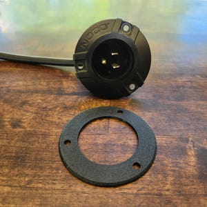 May include: A black NOCO brand AC port plug with an attached black power cord and a separate circular black mounting gasket featuring three screw holes. The plug has a round face with three metal prongs visible inside the central socket area.