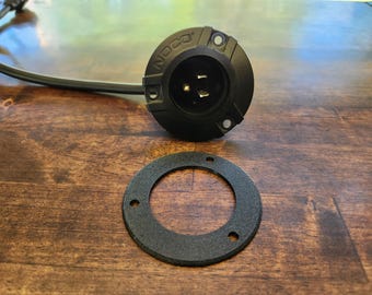 Könnte beinhalten: Ein schwarzer NOCO-Netzstecker mit angeschlossenem schwarzem Stromkabel und einer separaten runden schwarzen Montagedichtung mit drei Schraubenlöchern. Der Stecker hat eine runde Vorderseite mit drei Metallstiften in der zentralen Buchse.