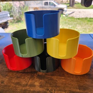 May include: A stack of six colorful cup holders in shades of blue, green, yellow, red, black, and orange. Each holder has a curved design with a slot for a handle. The holders are stacked on a wooden surface.