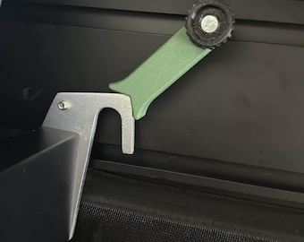 Könnte beinhalten: Nahaufnahme eines Verriegelungsmechanismus aus Metall und Kunststoff. Der Mechanismus besteht aus einer silberfarbenen Metallhalterung mit einem gebogenen Haken, einem grünen Kunststoffhebel und einem schwarzen Kunststoffknopf. Der Hintergrund ist ein dunkler, gesteppter Stoff.