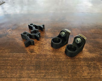 Könnte beinhalten: Draufsicht auf mehrere schwarze 3D-gedruckte Teile auf einer Holzoberfläche. Es gibt zwei Paare von Objekten: zwei kleine, gebogene Teile und zwei größere Teile mit kreisförmigen Öffnungen und Metallbolzen. Die Teile sind wahrscheinlich für mechanische Zwecke.