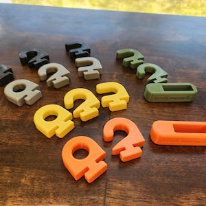 May include: A collection of colorful, 3D-printed objects, including the number "2" in various colors: black, gray, yellow, and orange. Also includes a green and orange rectangular object. The items are arranged on a wooden surface.