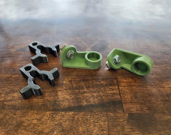 Könnte beinhalten: Eine Sammlung von 3D-gedruckten Hardwarekomponenten. Zwei grüne Teile mit kreisförmigen Öffnungen und angebrachten Schrauben werden zusammen mit drei schwarzen, stilisierten, bügelförmigen Teilen gezeigt. Die Artikel sind auf einer dunklen Holzmaserungsoberfläche angeordnet.