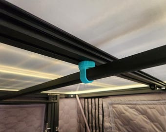 Könnte beinhalten: Nahaufnahme eines schwarzen Metallrahmens mit einem hellblauen Clip, der eine schwarze Stange sichert. Der Rahmen ist Teil einer größeren Struktur, mit einer weißen Decke und grauem Steppstoff im Hintergrund. Der Clip scheint aus Kunststoff zu sein.