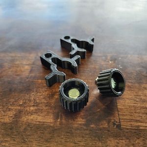 May include: A collection of black 3D-printed parts, including two gear-shaped pieces and two cylindrical components with internal details. The items are arranged on a wooden surface, showcasing their design and texture.