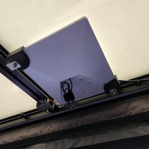 May include: A white rectangular electronic device mounted to a black metal frame with black plastic brackets. The device features a central cable connection, a coiled black power cord, and a small black component attached to the frame rail.