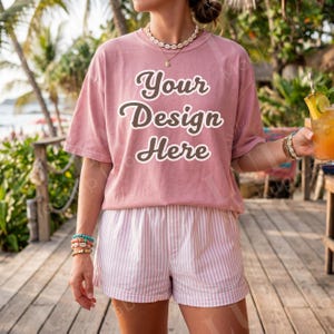 May include: A dusty rose t-shirt with the text "Your Design Here" in a script font. The model is wearing white and pink striped shorts and a shell necklace. The scene is set outdoors with a tropical vibe.