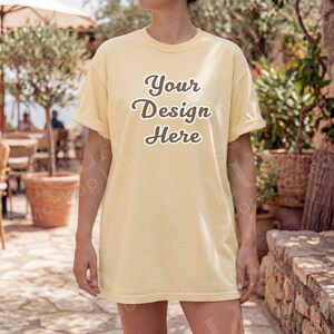 May include: A light yellow t-shirt with rolled-up sleeves. The text "Your Design Here" is printed on the front in a stylized font. The shirt is worn by a person outdoors.