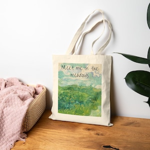 Könnte beinhalten: Beige Tragetasche mit Landschaftsmalerei und dem Text "Meet me in the meadows". Das Kunstwerk zeigt eine grüne Wiese unter blauem und weißem Himmel. Die Tasche hat zwei beige Griffe.
