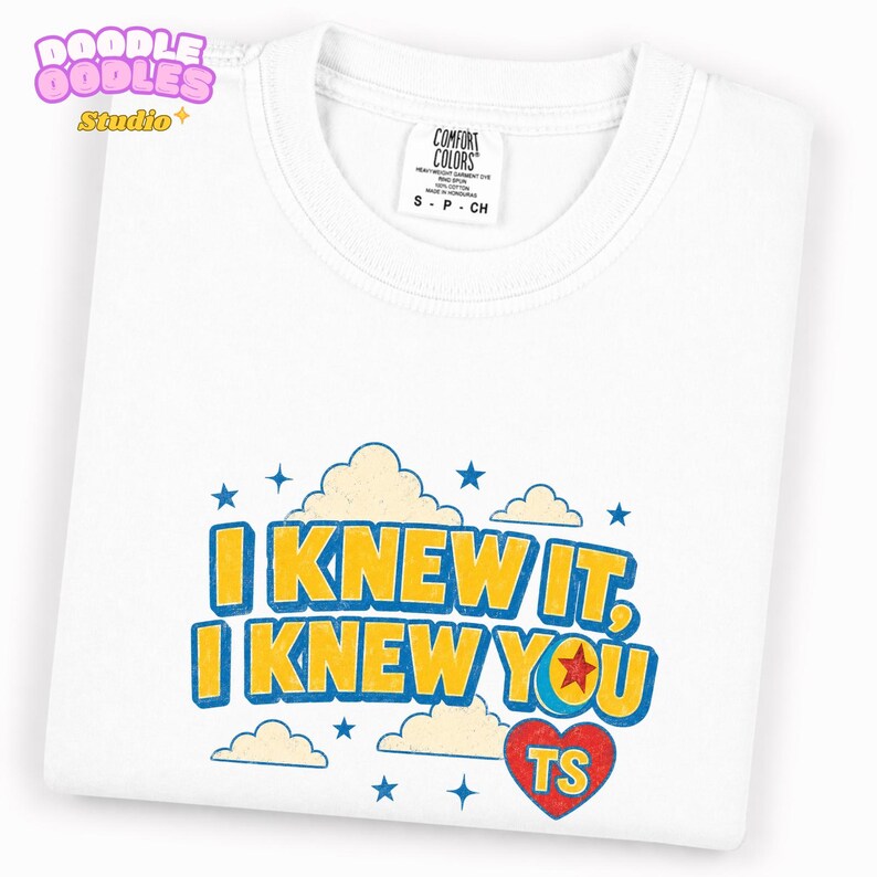 May include: A white short-sleeve t-shirt featuring a graphic print with the text I KNEW IT, I KNEW YOU in yellow letters with blue outlines. The design includes light yellow clouds, blue stars, and a red heart with the letters TS inside.