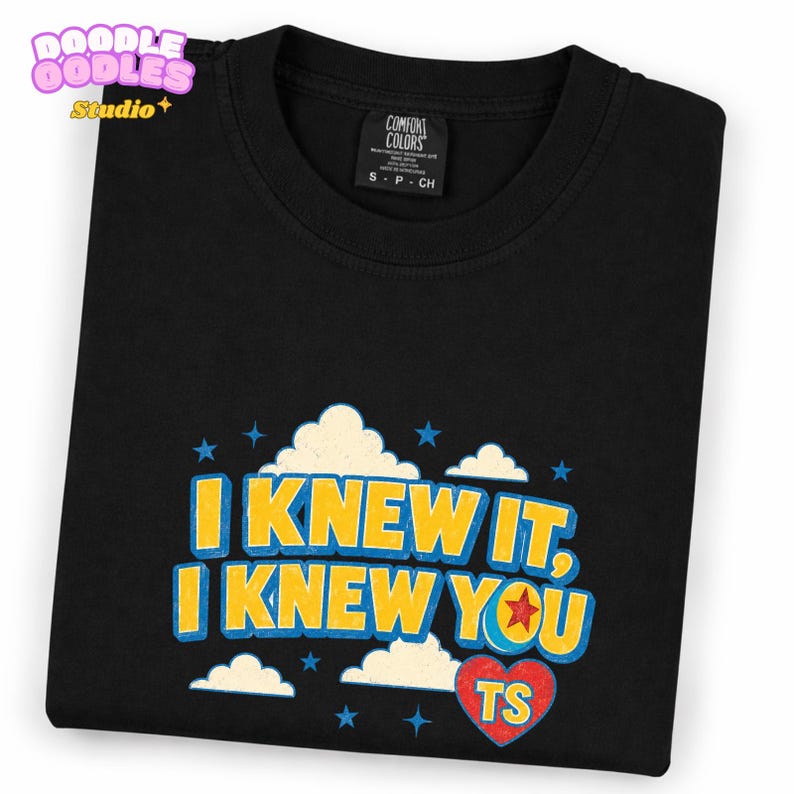 May include: A black t-shirt featuring a graphic print with the text I KNEW IT, I KNEW YOU in yellow letters with blue outlines. The design includes white clouds, blue stars, and a red heart with the letters TS inside, all set against a black fabric.