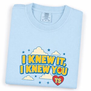 May include: A light blue t-shirt featuring a graphic print with yellow text that reads I KNEW IT, I KNEW YOU. The design includes cream-colored clouds, blue stars, and a red heart with the letters TS inside, all set against the light blue fabric.