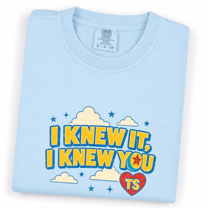 May include: A light blue t-shirt featuring a graphic print with yellow text that reads I KNEW IT, I KNEW YOU. The design includes cream-colored clouds, blue stars, and a red heart with the letters TS inside, all set against the light blue fabric.