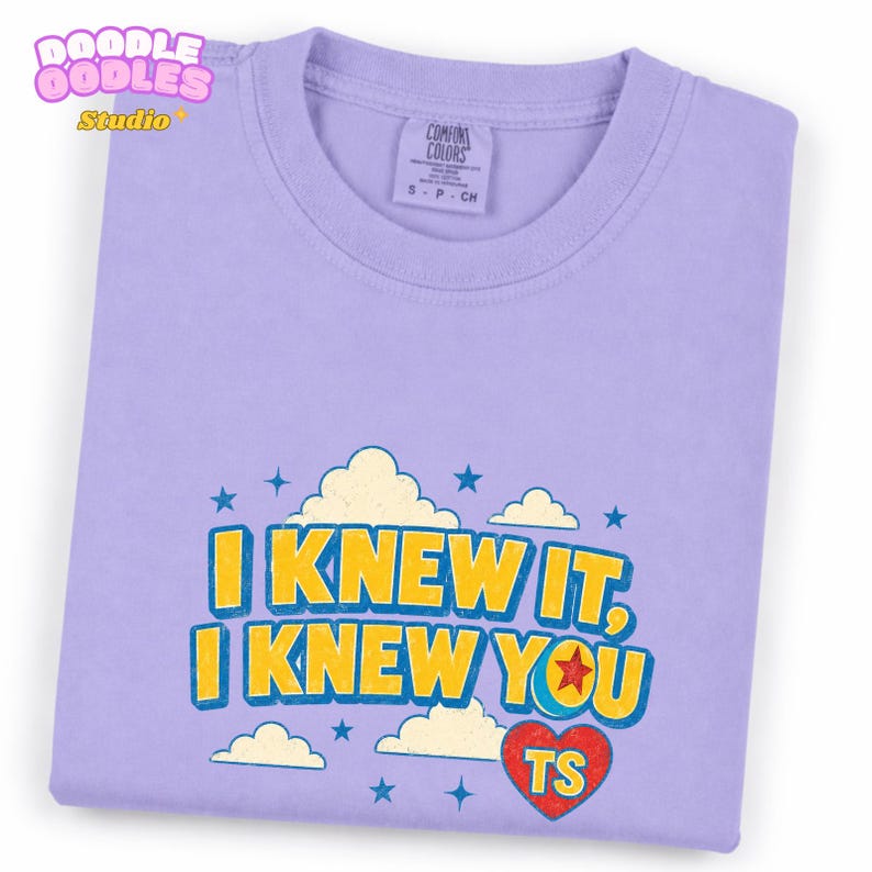 May include: A light purple t-shirt featuring a graphic print with yellow and blue text that reads I KNEW IT, I KNEW YOU. The design includes white clouds, blue stars, and a red heart with the letters TS inside, all set against the lavender fabric.