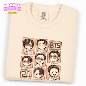 Peut inclure: Un t-shirt couleur crème arborant une impression graphique de sept personnages de style chibi dans des cadres carrés individuels, avec le texte "BTS" et "2.0". Le t-shirt présente un col rond et une étiquette Comfort Colors à l'intérieur.