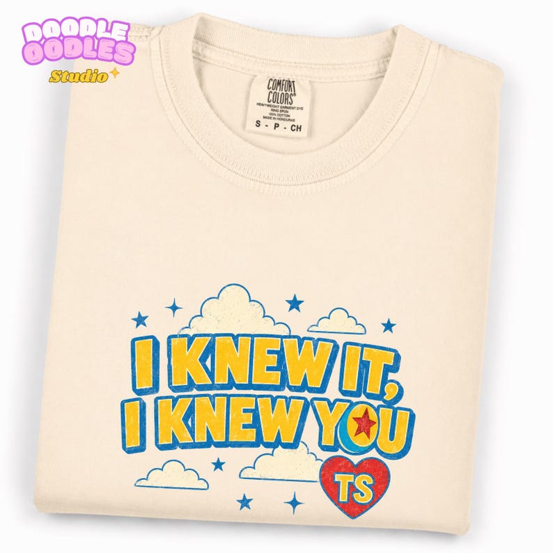 May include: A cream-colored t-shirt featuring a graphic print with blue clouds and stars. The text reads "I KNEW IT, I KNEW YOU" in yellow with blue outlines. A red heart contains the letters "TS" and a small red star inside the letter O.