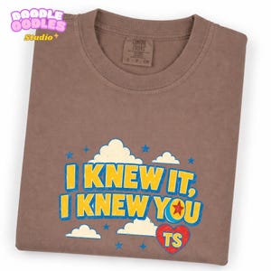 May include: A brown short-sleeved t-shirt featuring a graphic print with the text "I KNEW IT, I KNEW YOU" in yellow letters with a blue outline. The design includes white clouds, blue stars, and a red heart with the letters "TS" inside.