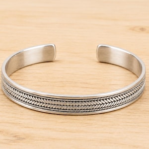 Könnte beinhalten: Ein silberfarbenes Armband in Manschettenform mit einem zentralen geflochtenen Seilmuster, das von zwei dünnen Perlenlinien flankiert wird. Das Armband hat eine glatte, polierte Innenseite und ein offenes Design mit einer detaillierten Metalloberfläche.