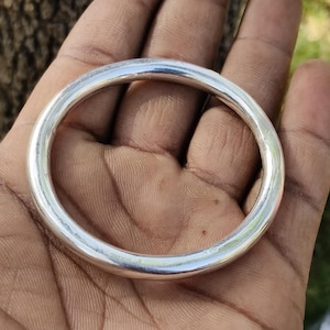 Puede incluir: Una pulsera tipo brazalete de tono plateado, lisa y pulida, con un diseño circular sencillo. La superficie metálica es reflectante y uniforme, con un perfil redondeado que crea un bucle continuo y sin costuras, sin cierres ni adornos.