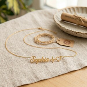 Op de afbeelding: Een goudkleurige ketting met de naam "Sophia" in cursief schrift, gevolgd door een hartslaglijn en een hartsymbool. De ketting ligt op een beige linnen oppervlak, met een opgerold certificaat op de achtergrond.