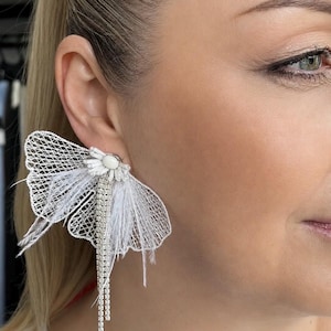May include: White, butterfly-shaped earrings with a delicate net design and feather accents. The earrings feature a central cluster of white beads and a long, dangling strand of sparkling rhinestones, creating a glamorous look.