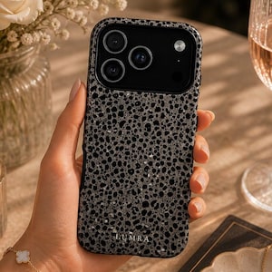 Op de afbeelding: Een smartphonehoesje met een textuur van zwarte en grijze spikkels en een matte afwerking. Het hoesje heeft een zwarte uitsparing voor de cameramodule en de merknaam LUMRA staat in kleine witte letters onderaan het achterpaneel.
