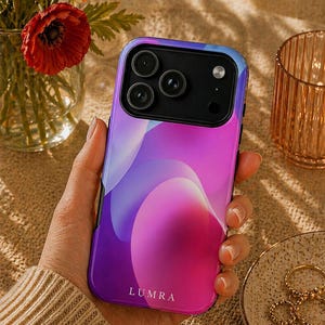 Peut inclure: Coque de smartphone avec un dégradé violet et rose vibrant. L'étui a une finition brillante et porte le nom de la marque "LUMRA" en bas. Il possède un module caméra noir avec trois objectifs.