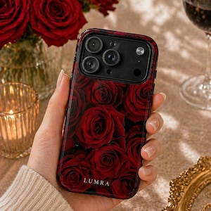 Op de afbeelding: Een smartphonehoesje met een patroon van rode rozen. De hoes heeft een zwarte rand en de merknaam "LUMRA" is onderaan zichtbaar. De hoes wordt in een hand gehouden, met een vage achtergrond met een vaas met rode rozen en een glas rode wijn.