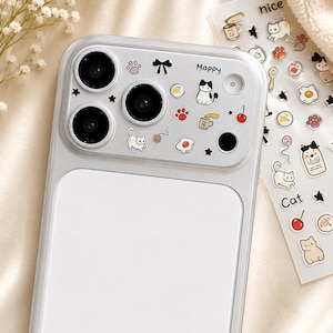 May include: A silver phone case featuring a camera lens protector decorated with small colourful stickers of cats, paw prints, stars, a bow, a coffee mug, and a fried egg, with the word Moppy printed near the top right corner of the lens area.