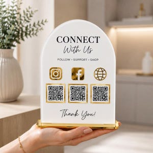 May include: An arched white acrylic sign with a gold base featuring the text CONNECT With Us, FOLLOW SUPPORT SHOP, and Thank You. The sign displays gold icons for Instagram, Facebook, and a website, each paired with a square QR code below it.