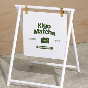 Può includere: Cartello bianco a cavalletto con la scritta "Kiyo Matcha" in verde. Il cartello include anche testo giapponese e una grafica di una catena montuosa. Il cartello è sostenuto da un telaio in metallo bianco con staffe color oro.