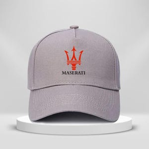 Op de afbeelding: Grijze baseballcap met het rode Maserati-logo. Het logo bevat een drietand boven de merknaam. De cap heeft een gebogen klep en een knoop aan de bovenkant.