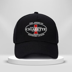Könnte beinhalten: Eine schwarze Baseballkappe mit einer weißen ovalen Grafik, die die rote Zahl 1 und den Text THE AMERICAN CIGARETTE RACING TEAM WORLD CHAMPION OFFSHORE LEGEND MADE IN USA in Weiß und Rot auf der Vorderseite der Kappe zeigt.