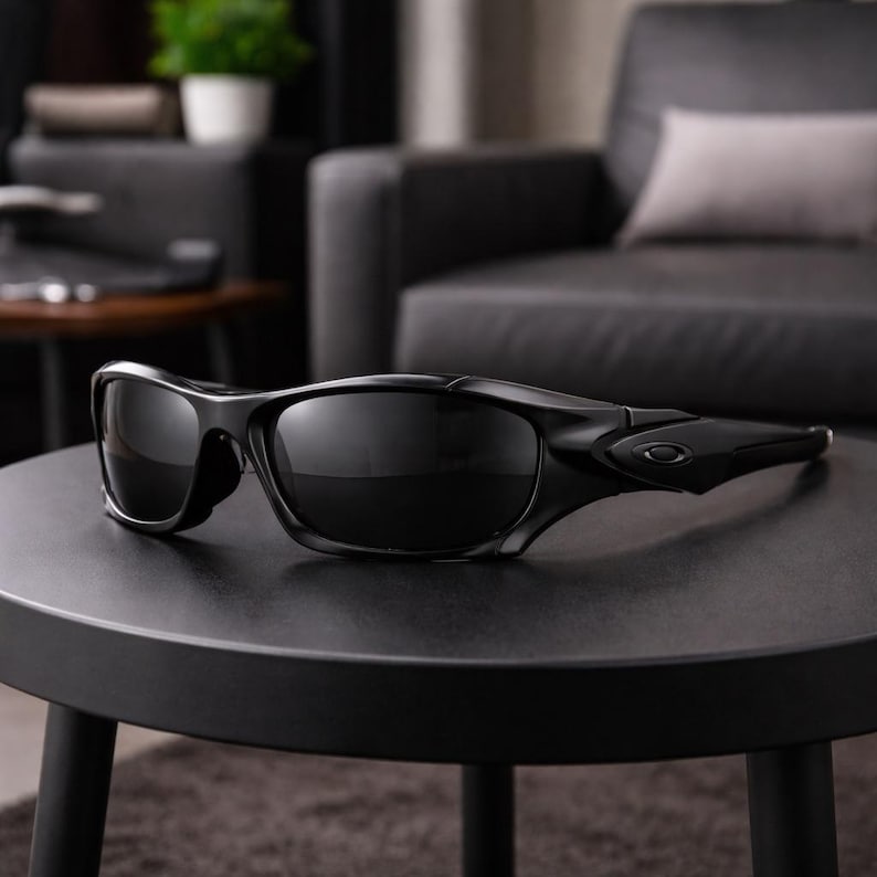 May include: Black sunglasses with a sleek, modern design. The sunglasses have a curved frame and dark lenses, resting on a black table. The sunglasses appear to be made of a durable material, suitable for outdoor activities.