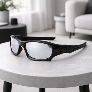 May include: Black sunglasses with silver mirrored lenses rest on a light gray table. The sunglasses have a sporty design with a thick black frame and curved temples. The lenses reflect the surrounding environment.