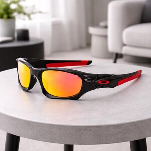 May include: Black sunglasses with red accents on the temples and orange-tinted lenses. The sunglasses are resting on a light gray surface. The frame is a glossy black, and the lenses have a gradient effect.