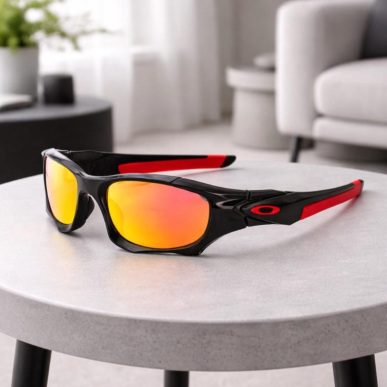 May include: Black sunglasses with red accents on the temples and orange-tinted lenses. The sunglasses are resting on a light gray surface. The frame is a glossy black, and the lenses have a gradient effect.