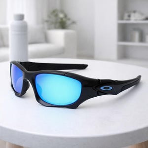 May include: Black sunglasses with blue mirrored lenses rest on a white surface. The sunglasses have a sporty design with a curved frame and the brand logo on the temple. The lenses reflect a bright blue color.