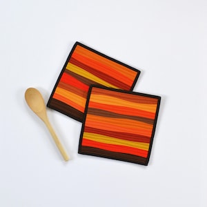 May include: Two square fabric pot holders featuring horizontal wavy stripes in shades of orange, yellow, red, and brown with black binding edges, accompanied by a single light-colored wooden spoon placed to the left of the quilted fabric items.